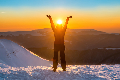 Person standing in snow with arms raised in front of rising sun.